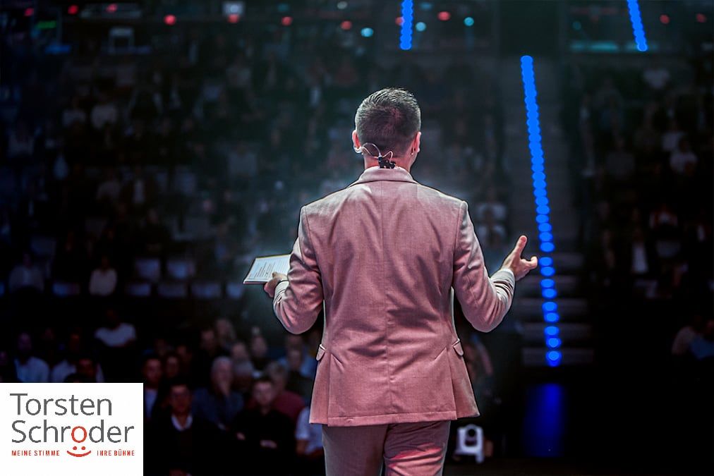 Keynote-Speaker von hinten auf der Bühne, Publikum im dunklen Saal mit blauer Akzentbeleuchtung