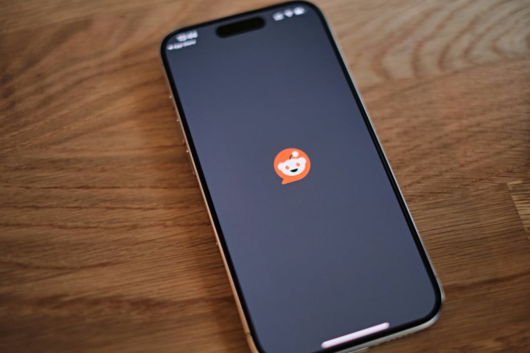 iPhone on a wooden desk showing the Reddit launch screen with the alien mascot logo